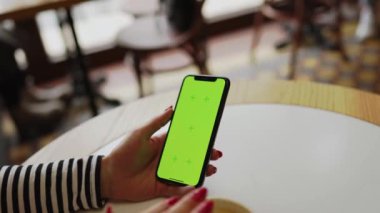 Genç bir kadın kahve dükkanında kahve içiyor ve telefonda çalışıyor. Kadın, telefon ekranını sarıyor. Telefonda yeşil ekran. Serbest konsept 4K