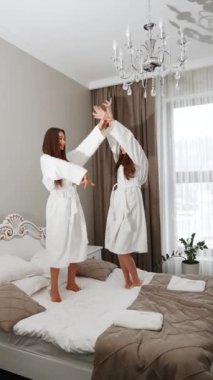 Bornozlu iki genç kadın yatakta dans edip kendilerini filme çekiyorlar. Otel tatili konsepti. Dikey video