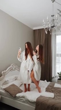 Bornozlu iki genç kadın yatakta dans edip kendilerini filme çekiyorlar. Otel tatili konsepti. Dikey video