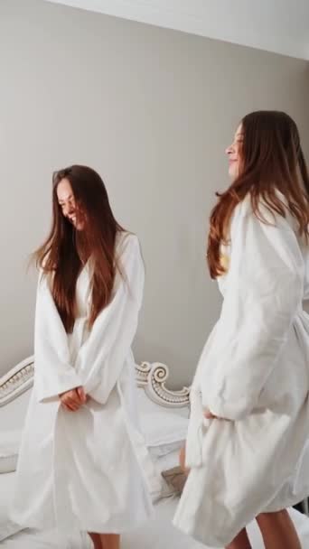 バスローブにいる2人の若い女性がベッドで踊り 自分自身を撮影します ホテルホリデーコンセプト バーティカルビデオ — ストック動画