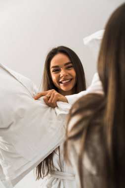 Bornozlu iki genç kadın yatakta gülüyor ve yastıklara vuruyor. Otel tatil konsepti