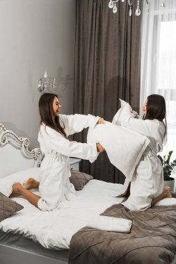 Bornozlu iki genç kadın yatakta gülüyor ve yastıklara vuruyor. Otel tatil konsepti