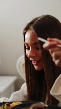 Bornozlu iki genç kadın yatakta kahvaltı yapıyor. Sağlıklı kahvaltı peynirli kekler. Otel tatili konsepti. Dikey video