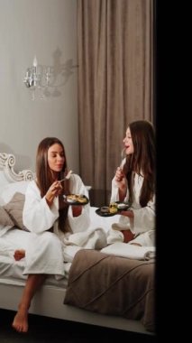 Bornozlu iki genç kadın yatakta kahvaltı yapıyor. Sağlıklı kahvaltı peynirli kekler. Otel tatili konsepti. Dikey video