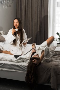 Bornozlu genç kadınlar yatakta kahvaltı yapar, kahve içer ve oynaşırlar. Kahvaltılık cheesecake ve meyveli kahve. Otel tatil konsepti