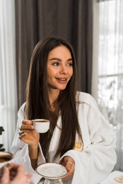 Bornozlu genç kadınlar yatakta kahvaltı yapar, kahve içer ve oynaşırlar. Kahvaltılık cheesecake ve meyveli kahve. Otel tatil konsepti