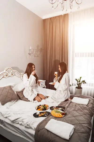 Young women in bathrobes have breakfast in bed, drink coffee and fool around. Breakfast cheesecakes and fruits with coffee. Hotel holiday concept