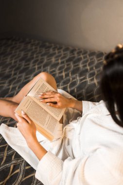 Bornozlu genç bir kadın yatakta kitap okuyor. Okuma ve eğitim kavramı..