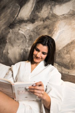 Bornozlu genç bir kadın yatakta kitap okuyor. Okuma ve eğitim kavramı..