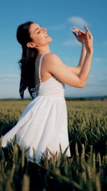 Beyaz elbiseli güzel bir kadın yeşil buğday tarlasında poz veriyor ve dans ediyor. Özgürlük kavramı. Dikey video