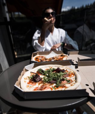 Bornozlu bir kadın dağa tepeden bakan bir balkonda pizza yiyor. Otel tatili konsepti. Pizza.