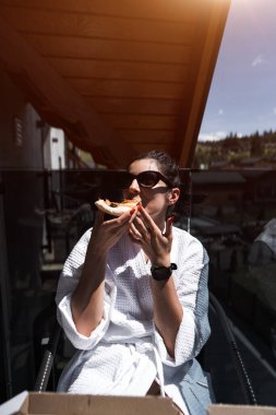 Bornozlu bir kadın dağa tepeden bakan bir balkonda pizza yiyor. Otel tatili konsepti. Pizza.