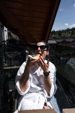 Bornozlu bir kadın dağa tepeden bakan bir balkonda pizza yiyor. Otel tatili konsepti. Pizza.