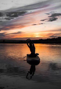 Genç bir kadın gün batımında gölde SUP sörf tahtasında yüzüyor. Gün batımında SUP tahtasında oturan bir kadının silüeti. SUP sörf konsepti