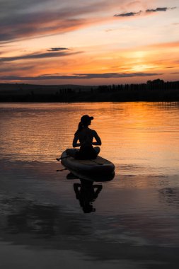 Genç bir kadın gün batımında gölde SUP sörf tahtasında yüzüyor. Gün batımında SUP tahtasında oturan bir kadının silüeti. SUP sörf konsepti