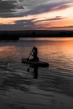 Genç bir kadın gün batımında gölde SUP sörf tahtasında yüzüyor. Gün batımında SUP tahtasında oturan bir kadının silüeti. SUP sörf konsepti