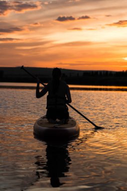 Genç bir kadın gün batımında gölde SUP sörf tahtasında yüzüyor. Gün batımında SUP tahtasında oturan bir kadının silüeti. SUP sörf konsepti