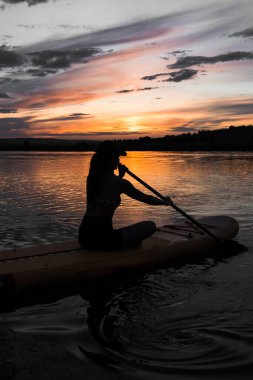 Genç bir kadın gün batımında gölde SUP sörf tahtasında yüzüyor. Gün batımında SUP tahtasında oturan bir kadının silüeti. SUP sörf konsepti