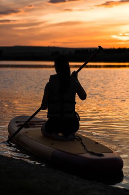 Genç bir kadın gün batımında gölde SUP sörf tahtasında yüzüyor. Gün batımında SUP tahtasında oturan bir kadının silüeti. SUP sörf konsepti