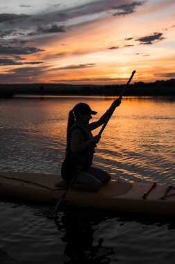 Genç bir kadın gün batımında gölde SUP sörf tahtasında yüzüyor. Gün batımında SUP tahtasında oturan bir kadının silüeti. SUP sörf konsepti