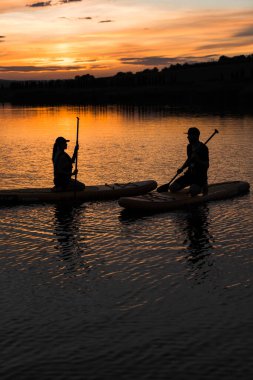 Bir adam ve bir kadın gün batımında bir gölde SUP tahtalarında yüzüyorlar. Gün batımında güneş ışınlarında sörf tahtasında bir erkek ve bir kadının siluetleri. SUP sörf konsepti