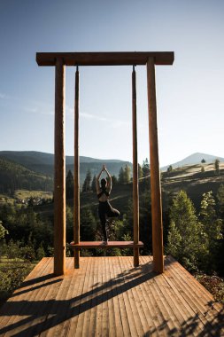 Genç bir kadın, dağların arka planında salıncakta yoga yapıyor. Yoga ve meditasyon konsepti