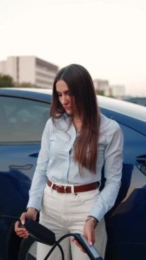 Genç bir kadın bir süpermarketin yakınındaki şarj istasyonunda bir arabayı şarj ediyor. Elektrikli bir arabayı şarj etmek. Elektrikli araba konsepti. Dikey video