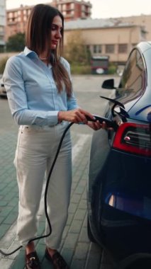 Genç bir kadın bir süpermarketin yakınındaki şarj istasyonunda bir arabayı şarj ediyor. Elektrikli bir arabayı şarj etmek. Elektrikli araba konsepti. Dikey video