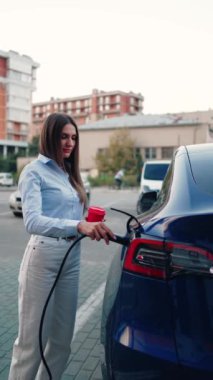 Genç bir kadın bir süpermarketin yakınındaki şarj istasyonunda bir arabayı şarj ediyor. Elektrikli bir arabayı şarj etmek. Elektrikli araba konsepti. Dikey video