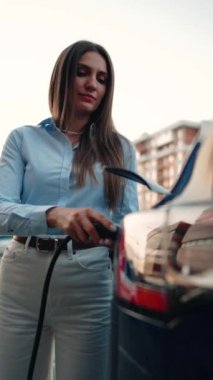 Genç bir kadın bir süpermarketin yakınındaki şarj istasyonunda bir arabayı şarj ediyor. Elektrikli bir arabayı şarj etmek. Elektrikli araba konsepti. Dikey video