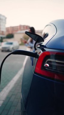 Genç bir kadın bir süpermarketin yakınındaki şarj istasyonunda bir arabayı şarj ediyor. Elektrikli bir arabayı şarj etmek. Elektrikli araba konsepti. Dikey video