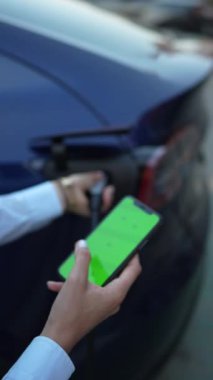 Genç bir kadın bir süpermarketin yakınındaki şarj istasyonunda bir arabayı şarj ediyor. Elektrikli bir arabayı şarj etmek. Elektrikli araba konsepti. Dikey video