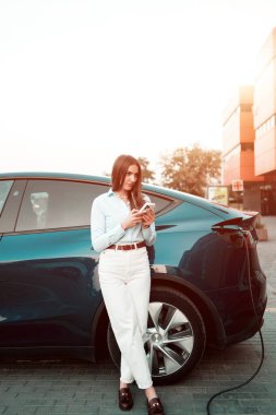 Genç bir kadın bir süpermarketin yakınındaki şarj istasyonunda bir arabayı şarj ediyor. Elektrikli bir arabayı şarj etmek. Elektrikli araba konsepti. Dikey fotoğraf