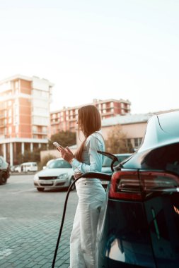Genç bir kadın bir süpermarketin yakınındaki şarj istasyonunda bir arabayı şarj ediyor. Elektrikli bir arabayı şarj etmek. Elektrikli araba konsepti. Dikey fotoğraf