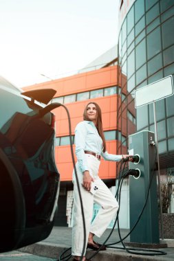 Genç bir kadın bir süpermarketin yakınındaki şarj istasyonunda bir arabayı şarj ediyor. Elektrikli bir arabayı şarj etmek. Elektrikli araba konsepti. Dikey fotoğraf