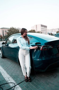 Genç bir kadın bir süpermarketin yakınındaki şarj istasyonunda bir arabayı şarj ediyor. Elektrikli bir arabayı şarj etmek. Elektrikli araba konsepti. Dikey fotoğraf