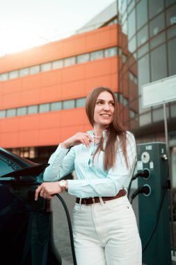 Genç bir kadın bir süpermarketin yakınındaki şarj istasyonunda bir arabayı şarj ediyor. Elektrikli bir arabayı şarj etmek. Elektrikli araba konsepti. Dikey fotoğraf