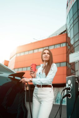 Genç bir kadın bir süpermarketin yakınındaki şarj istasyonunda bir arabayı şarj ediyor. Elektrikli bir arabayı şarj etmek. Elektrikli araba konsepti. Dikey fotoğraf