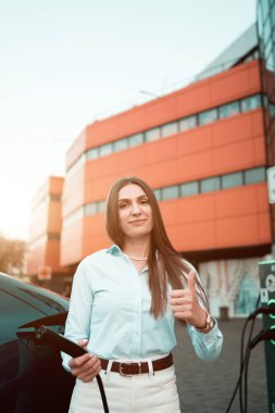 Genç bir kadın bir süpermarketin yakınındaki şarj istasyonunda bir arabayı şarj ediyor. Elektrikli bir arabayı şarj etmek. Elektrikli araba konsepti. Dikey fotoğraf