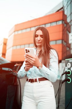 Genç bir kadın bir süpermarketin yakınındaki şarj istasyonunda bir arabayı şarj ediyor. Elektrikli bir arabayı şarj etmek. Elektrikli araba konsepti. Dikey fotoğraf
