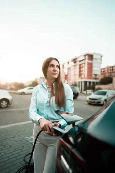Genç bir kadın bir süpermarketin yakınındaki şarj istasyonunda bir arabayı şarj ediyor. Elektrikli bir arabayı şarj etmek. Elektrikli araba konsepti. Dikey fotoğraf