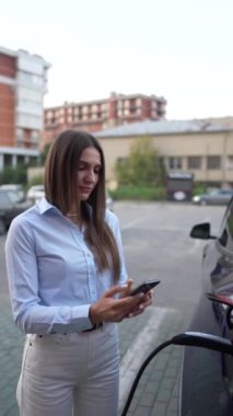 Genç bir kadın bir süpermarketin yakınındaki şarj istasyonunda bir arabayı şarj ediyor. Elektrikli bir arabayı şarj etmek. Elektrikli araba konsepti. Dikey video
