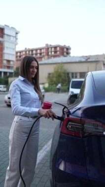 Genç bir kadın bir süpermarketin yakınındaki şarj istasyonunda bir arabayı şarj ediyor. Elektrikli bir arabayı şarj etmek. Elektrikli araba konsepti. Dikey video