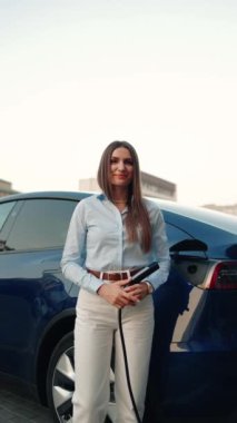 Genç bir kadın bir süpermarketin yakınındaki şarj istasyonunda bir arabayı şarj ediyor. Elektrikli bir arabayı şarj etmek. Elektrikli araba konsepti. Dikey video