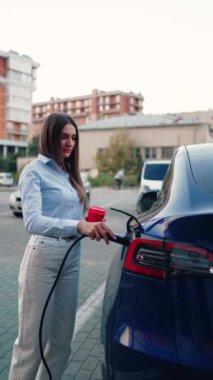 Genç bir kadın bir süpermarketin yakınındaki şarj istasyonunda bir arabayı şarj ediyor. Elektrikli bir arabayı şarj etmek. Elektrikli araba konsepti. Dikey video