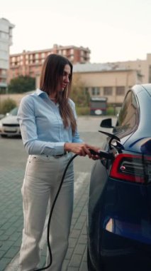 Genç bir kadın bir süpermarketin yakınındaki şarj istasyonunda bir arabayı şarj ediyor. Elektrikli bir arabayı şarj etmek. Elektrikli araba konsepti. Dikey video