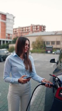 Genç bir kadın bir süpermarketin yakınındaki şarj istasyonunda bir arabayı şarj ediyor. Elektrikli bir arabayı şarj etmek. Elektrikli araba konsepti. Dikey video