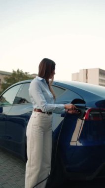 Genç bir kadın bir süpermarketin yakınındaki şarj istasyonunda bir arabayı şarj ediyor. Elektrikli bir arabayı şarj etmek. Elektrikli araba konsepti. Dikey video