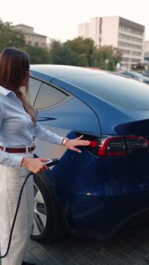 Genç bir kadın bir süpermarketin yakınındaki şarj istasyonunda bir arabayı şarj ediyor. Elektrikli bir arabayı şarj etmek. Elektrikli araba konsepti. Dikey video