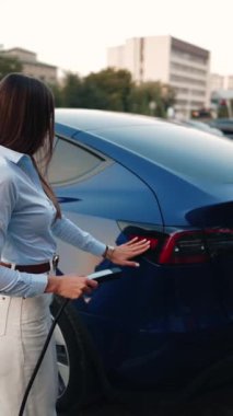 Genç bir kadın bir süpermarketin yakınındaki şarj istasyonunda bir arabayı şarj ediyor. Elektrikli bir arabayı şarj etmek. Elektrikli araba konsepti. Dikey video